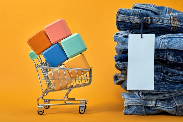 Uma pilha de jeans azuis com uma etiqueta branca em branco em um fundo amarelo. Carrinho de compras com cubos multicoloridos. Vendas realizadas — Fotografia de Stock