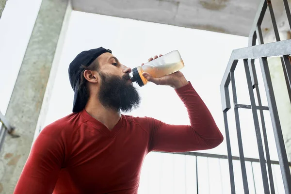 Vista lateral del deportista barbudo activo bebiendo agua de la botella de vidrio mientras descansa y se sienta en las escaleras en un día brillante — Foto de Stock