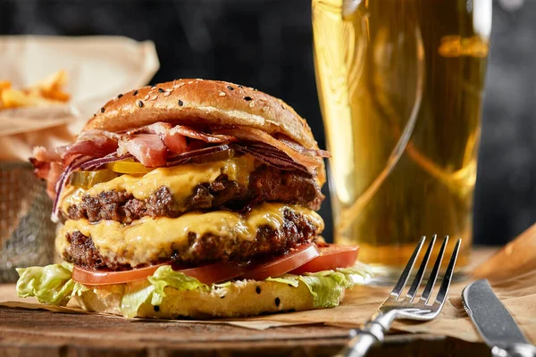 Koncept amerického fast foodu. Šťavnatý americký burger se dvěma hovězími placičkami a tmavým pivem na skleněném stole na dřevěném pozadí. Kopírovat prostor — Stock fotografie
