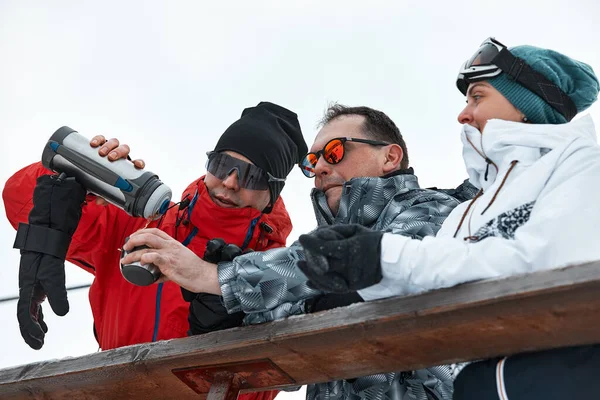 Gruppo di sciatori amici sulla montagna stanno riposando e bevendo caffè da un thermos sullo sfondo degli impianti di risalita — Foto Stock