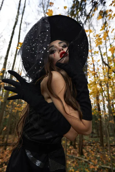 Jeune femme en robe noire avec chapeau de sorcière et citrouille orange placée dans la forêt, concept Halloween. Thème d'horreur. — Photo