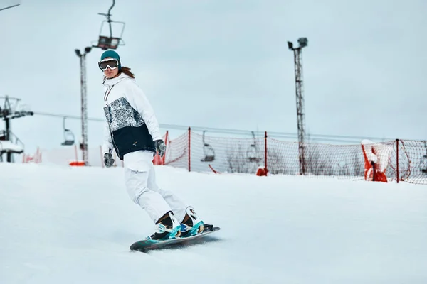 Fröhliche Frau fährt im Winter auf dem Snowboard in den Bergen. Frau im weißen Skianzug — Stockfoto