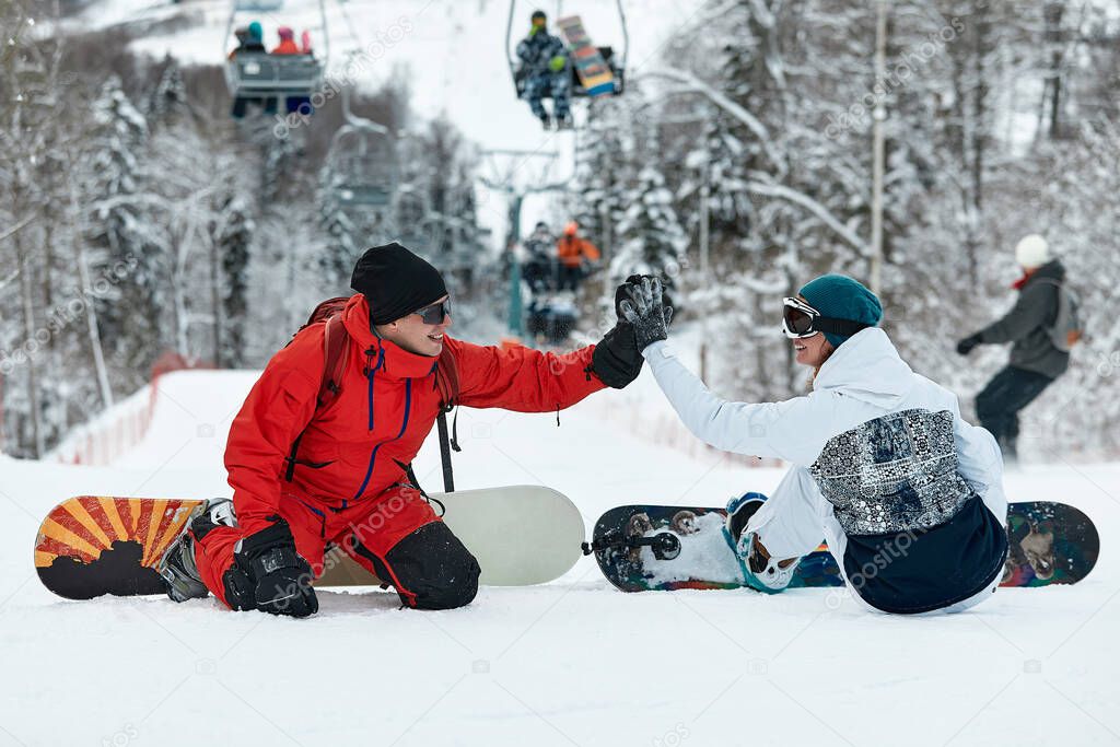 Couple of snowboarders gives a high five to each other while sitting on the snow tops of a slope against a blue sky and ski lift on the background at winter ski resort