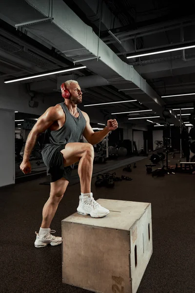 Sexy gespierde mannen met behulp van het platform voor zijn benen op een donkere kleurrijke achtergrond van gym. — Stockfoto