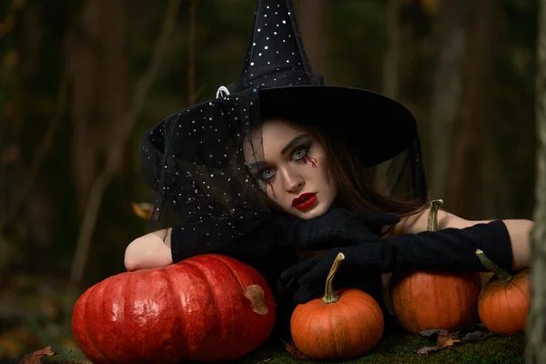 Mujer joven en vestido negro con sombrero de bruja y calabaza naranja colocada en el bosque, concepto de Halloween. Tema Horror. — Foto de Stock