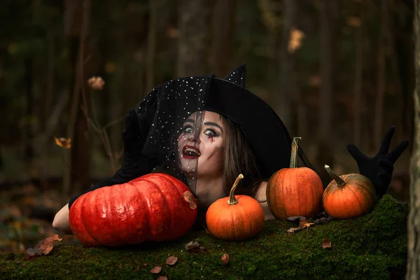 Jeune femme en robe noire avec chapeau de sorcière et citrouille orange placée dans la forêt, concept Halloween. Thème d'horreur. — Photo