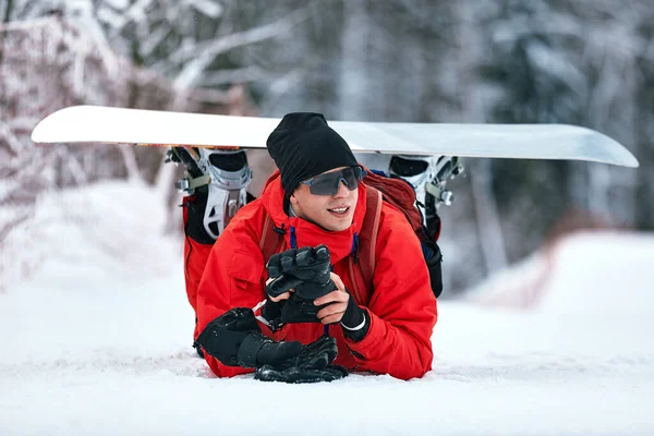 Hombre en traje de esquí rojo se sienta en la colina con su tabla de snowboard — Foto de Stock