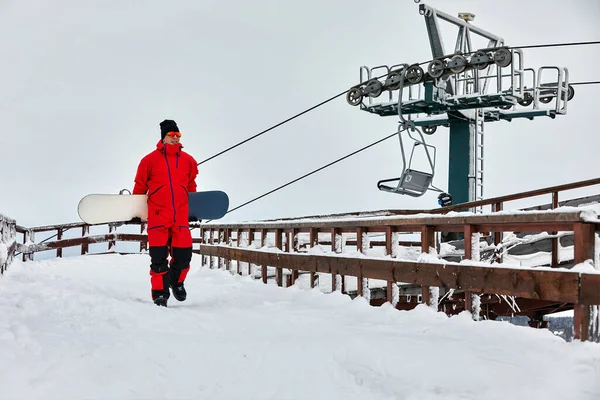 Чоловічий сноубордист у червоному костюмі, що йде на засніженому пагорбі зі сноубордом, концепцією катання на лижах та сноуборді — стокове фото
