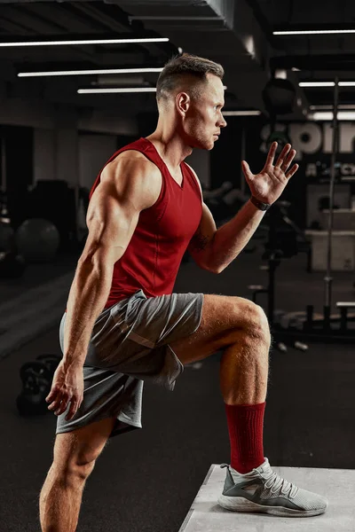 Sexy gespierde mannen met behulp van het platform voor zijn benen op een donkere kleurrijke achtergrond van gym. — Stockfoto