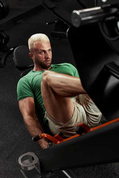 Een man, een bodybuilder doet oefeningen op zijn benen. De atleet traint zijn benen in de sportschool in de simulator. — Stockfoto