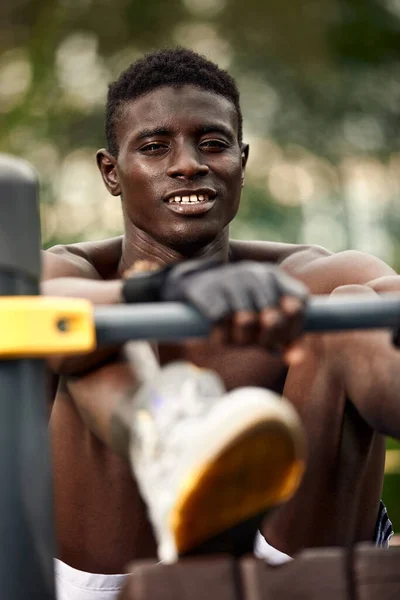 A smiling African American athlete performs abdominal exercises, an athlete in excellent physical shape does abdominal exercises on the street in the sports field. — Foto de Stock