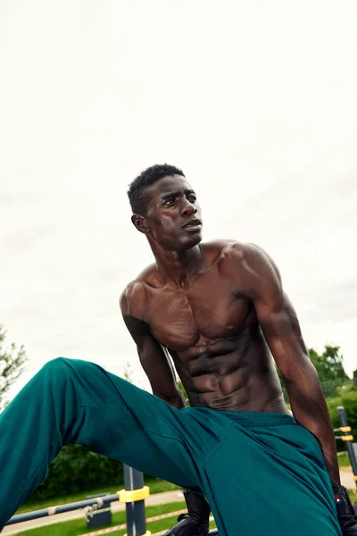 Joven musculoso ejercitándose en el campo de deportes. Hombre africano mirando hacia un lado mientras hace ejercicios de barra horizontal. Modelo masculino sin camisa que hace ejercicio al aire libre. — Foto de Stock