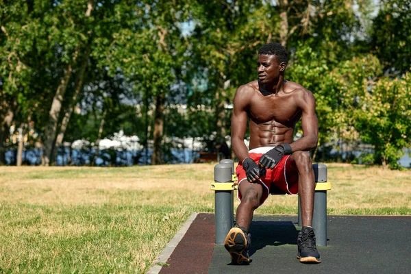 Joven musculoso ejercitándose en el campo de deportes. Hombre africano mirando hacia un lado mientras hace ejercicios de barra horizontal. Modelo masculino sin camisa que hace ejercicio al aire libre. — Foto de Stock