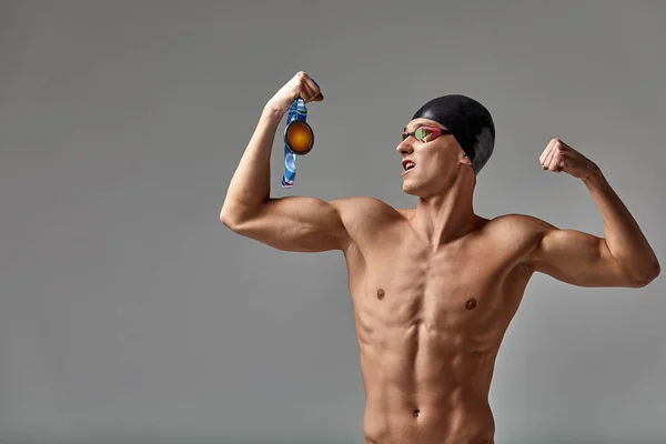 Nadador com uma medalha em um fundo cinza se alegra na vitória, um atleta em excelente forma física celebra uma vitória segurando uma medalha em sua mão, conceito de vitória, fundo cinza, espaço de cópia — Fotografia de Stock