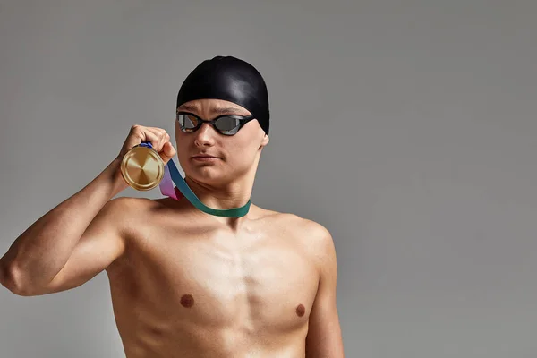 Nadador com uma medalha em um fundo cinza se alegra na vitória, um atleta em excelente forma física celebra uma vitória segurando uma medalha em sua mão, conceito de vitória, fundo cinza, espaço de cópia — Fotografia de Stock