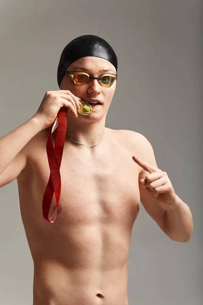 Nadador con una medalla sobre un fondo gris se regocija en la victoria, un atleta en excelente forma física celebra una victoria agarrando una medalla en su mano, concepto de victoria, fondo gris, espacio de copia — Foto de Stock