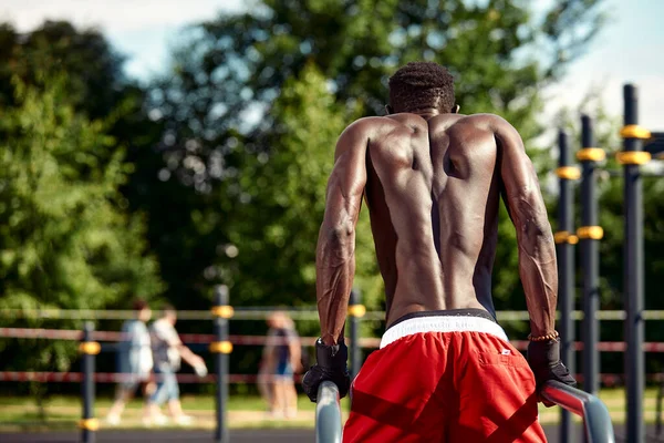 Atleta afroamericano musculoso con un torso esculpido desnudo haciendo flexiones en las barras irregulares en el paisaje urbano, disparo de espalda, espacio de copia — Foto de Stock