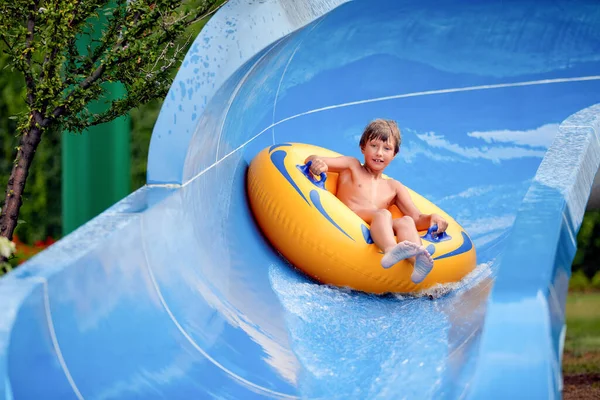 Feliz um menino de 8 anos está montando no parque aquático em círculos infláveis em escorregas de água com respingo — Fotografia de Stock