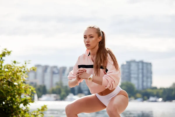 Blonde young athletic woman doing squats in a park in an urban environment. Attractive athletic woman exercising outdoors in the morning, copy space. Health, fitness concept