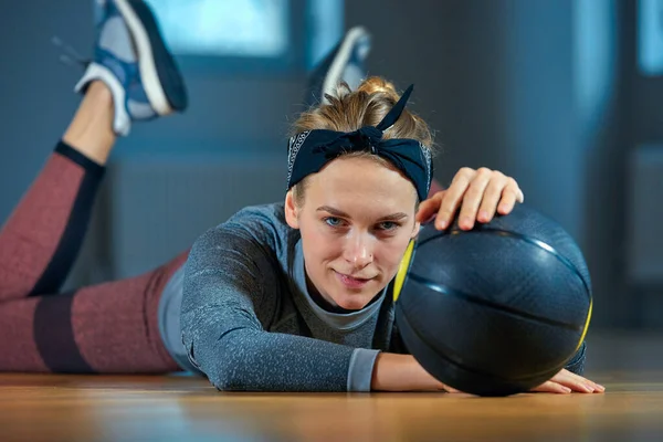 Smuk pasform kvinde i sportstøj poserende mens liggende på gulvet med basketball foran vinduet på gym Sund pige livsstil og sport koncept - Stock-foto