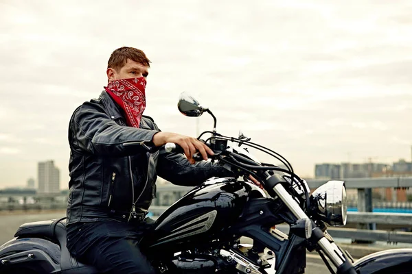 Porträtt av snygg man klädd i en svart läderjacka och bandana sitter på sin skräddarsydda retro motorcykel, tittar bort, byggnader i storstaden på bakgrunden. — Stockfoto