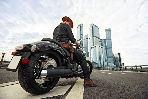 Porträtt av snygg man klädd i en svart läderjacka och bandana med pistol sitter på sin skräddarsydda retro motorcykel, tittar bort, byggnader i storstaden på bakgrunden. — Stockfoto