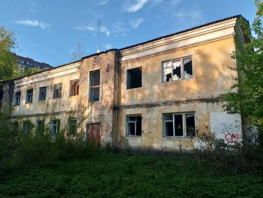 An abandoned building with peeling yellow paint and broken windows. Overgrown vegetation surrounds the structure, indicating years of neglect.