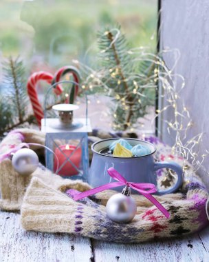 Noel süslemeleri, kremalı bir fincan kahve, yanan mumlu bir fener, ladin dalları ve ahşap bir masada sıcak bir eşarp, ev konforu, tebrikler. 