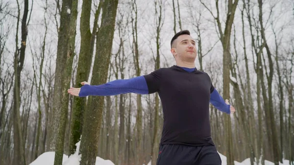 The athlete does exercises in the winter forest. Athlete training in winter forest, trail runner, fit man doing exercises in cold weather. A guy in a sports uniform is doing exercises.