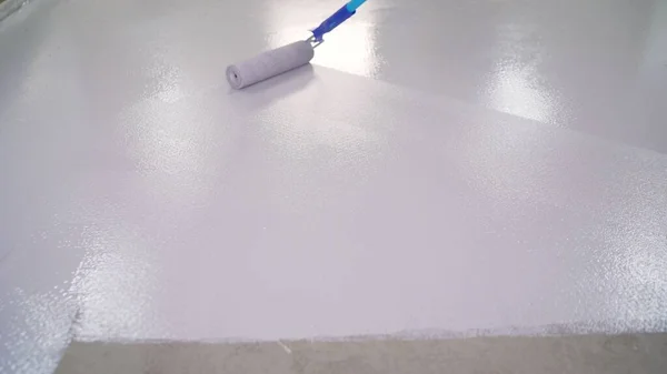A worker paints the floor white with a roller. The concrete floor is painted with white paint. In focus Paint roller with defocused background. floor painting. dirty repairs and alterations.