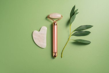 Gua sha aracı ve yüz masajı silindiri yeşil arka planda, güzellik soyut fotoğrafı. Üst görünüm
