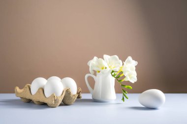 Paskalya yumurtaları ve çiçekler. Karton kutuda beyaz yumurtalar ve seramik sürahide Freesia beyaz bahar buketi..