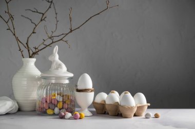 Kavanozda mini yumurta şekerlemeleri, vazoda dallar ve beyaz tavuk yumurtaları olan paskalya arkaplanı.