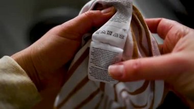 Çamaşır makinesinin yanındaki giysi hakkında dikkatli semboller ve bilgi içeren etiket okuyan kadın, yakın plan
