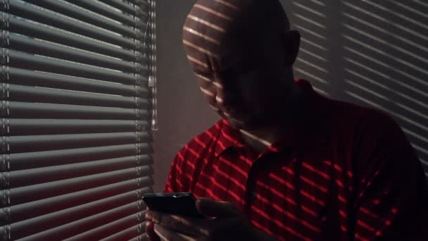 Мужчина со смартфоном стоит у окна с жалюзи — стоковое видео