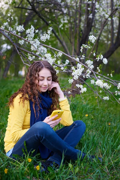 Bir akıllı telefon ile çimenlerin üzerinde oturan güzel genç kadın — Stok fotoğraf