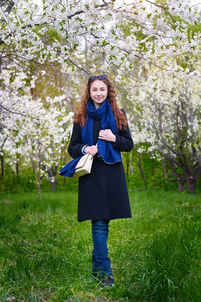 Young woman in coat walking at blossoming spring cherry garden — Stock Photo, Image