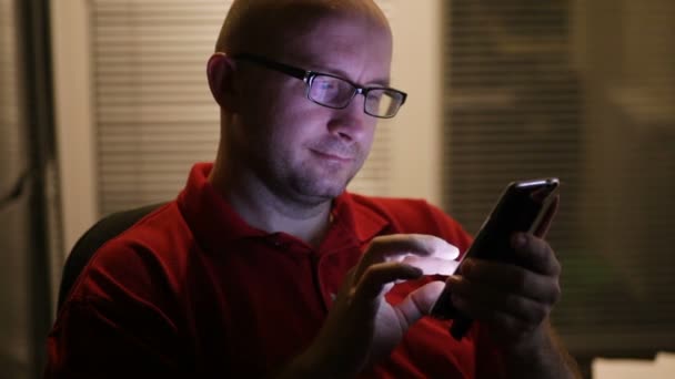 Ο άνθρωπος με τα γυαλιά που κάθεται σε ένα γραφείο, χρησιμοποιώντας ένα smartphone — Αρχείο Βίντεο