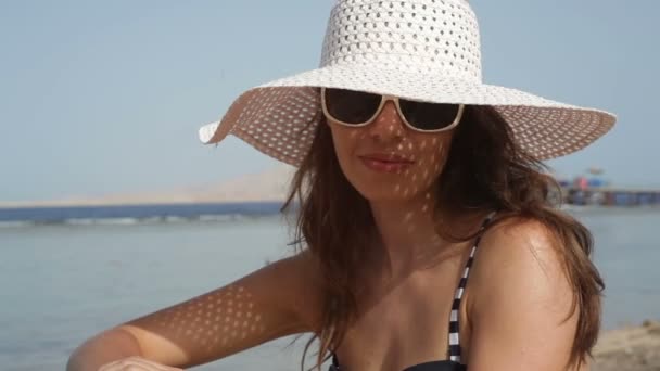 Молодая женщина в очках и шляпе на пляже у моря — стоковое видео