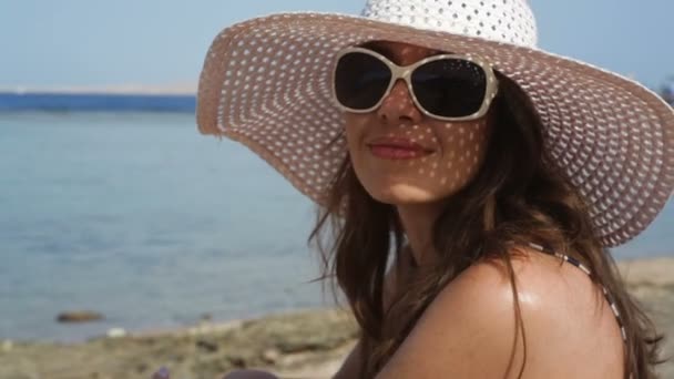 Ung kvinna i glas och en hatt på en strand nära havet — Stockvideo