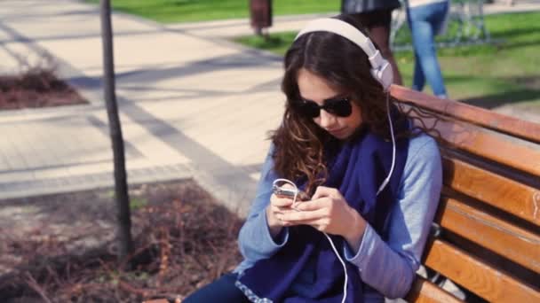 ベンチの上の公園でヘッドフォンで音楽を聴くサングラスの若い女性 — ストック動画