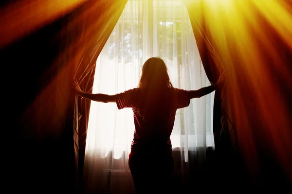 Silhouette of a young woman opens curtains at window — Stock Photo, Image