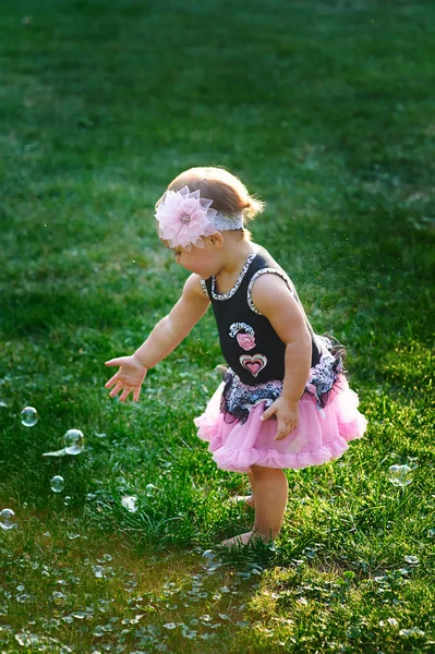 Holčička v růžových šatech fouká mýdlové bubliny na letní louku na slunečný den, pohled zezadu — Stock fotografie