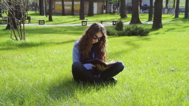Mooie jonge vrouw in het park op het gras zitten en lezen van een boek — Stockvideo