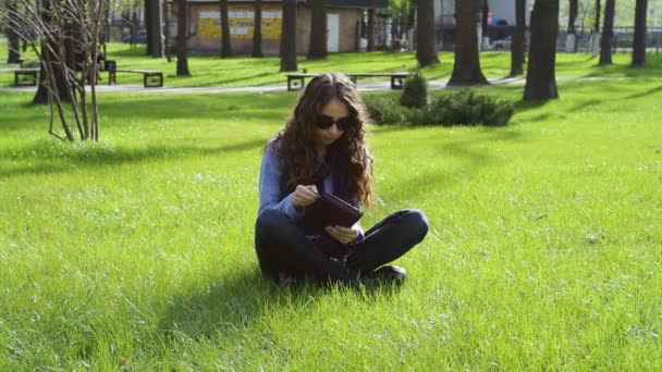 Hermosa joven sentada en el parque en la hierba y leyendo un libro — Vídeos de Stock