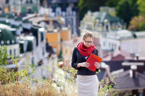 Junge Frau mit Brille und roter Mappe in der Hand vor dem Hintergrund der Stadt — Stockfoto