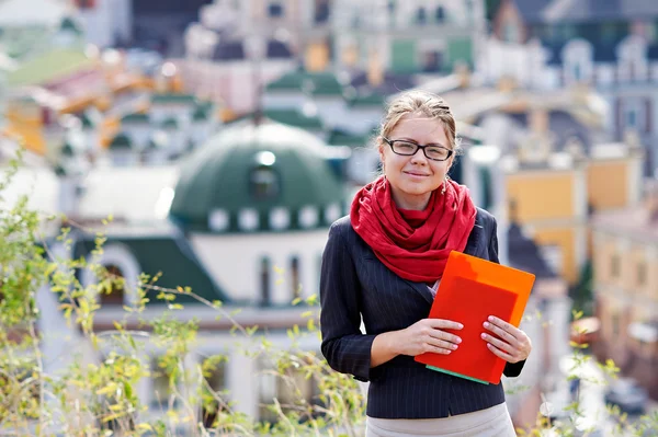 Mujer joven en gafas con carpeta roja en las manos sobre el fondo de la ciudad — Foto de Stock