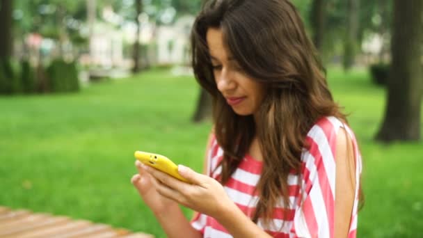 Mujer sonriente joven usando teléfono inteligente sentado en el banco en el parque — Vídeo de stock