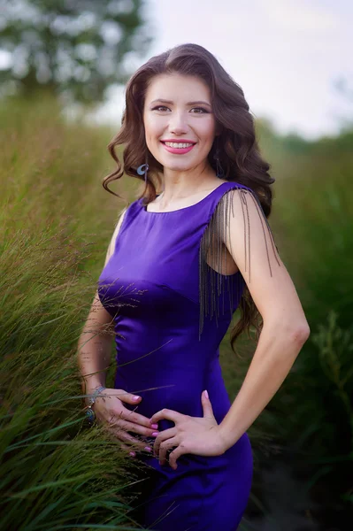 若いきれいな緑の芝生の上に立ってエレガントな紫のドレスで髪の長い風が強い女性 — ストック写真