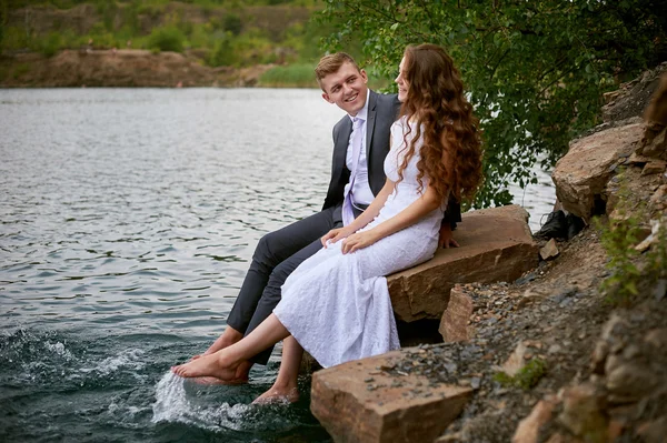 Loving wedding couple sitting and kissing near water — Stock Photo, Image